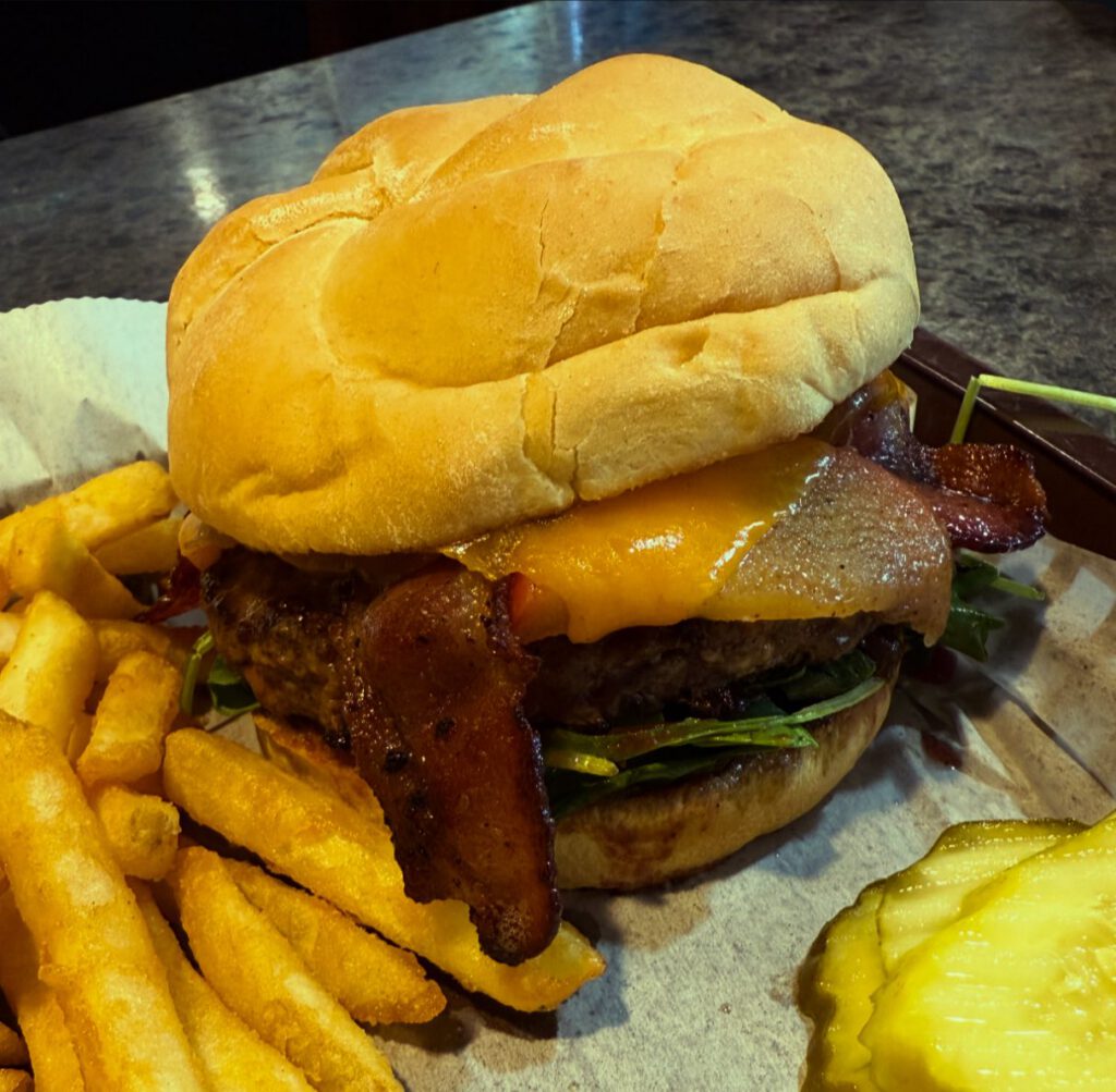 Burger of the Month ‘How ’Bout Dem Apples’ featuring a half-pound beef patty with bacon, melted cheddar, arugula, apple butter BBQ sauce, and brandied apples, served with fries and a pickle.