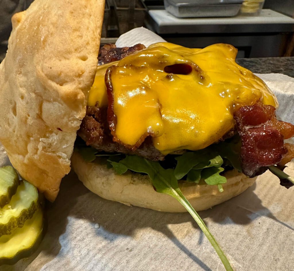 Wiso Jam Burger with a half-pound beef patty topped with melted cheddar cheese, bacon, arugula, and house-made brandy old-fashioned jam on a toasted bun with a pickle on the side.
