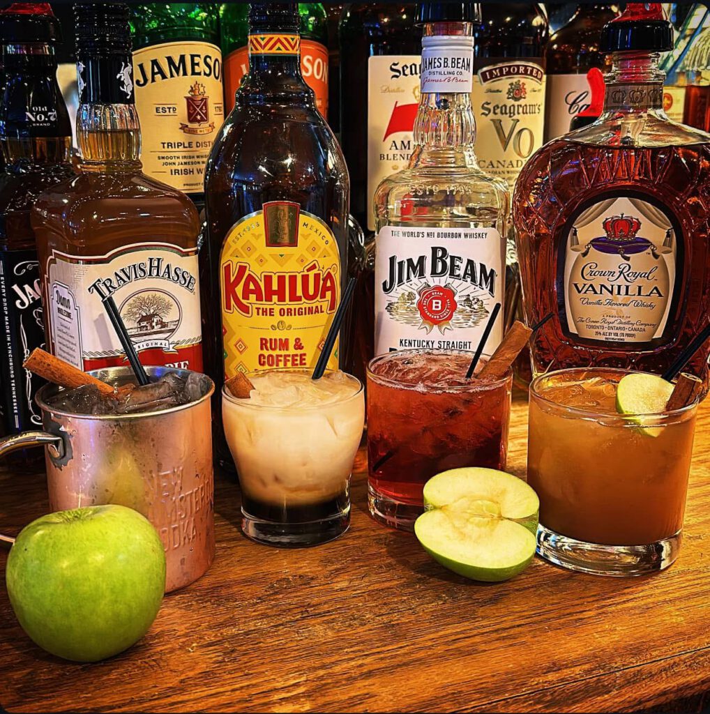 Seasonal cocktail display featuring Apple Butter Old Fashioned, Cranberry Cobbler Smash, Pumpkin Spice White Russian, and Apple Pie Mule, shown with liquor bottles, apples, and mixed drinks on a bar.
